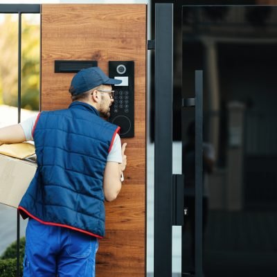 Back view of a deliverer ringing on intercom at gate of a customer's house while delivering packages.